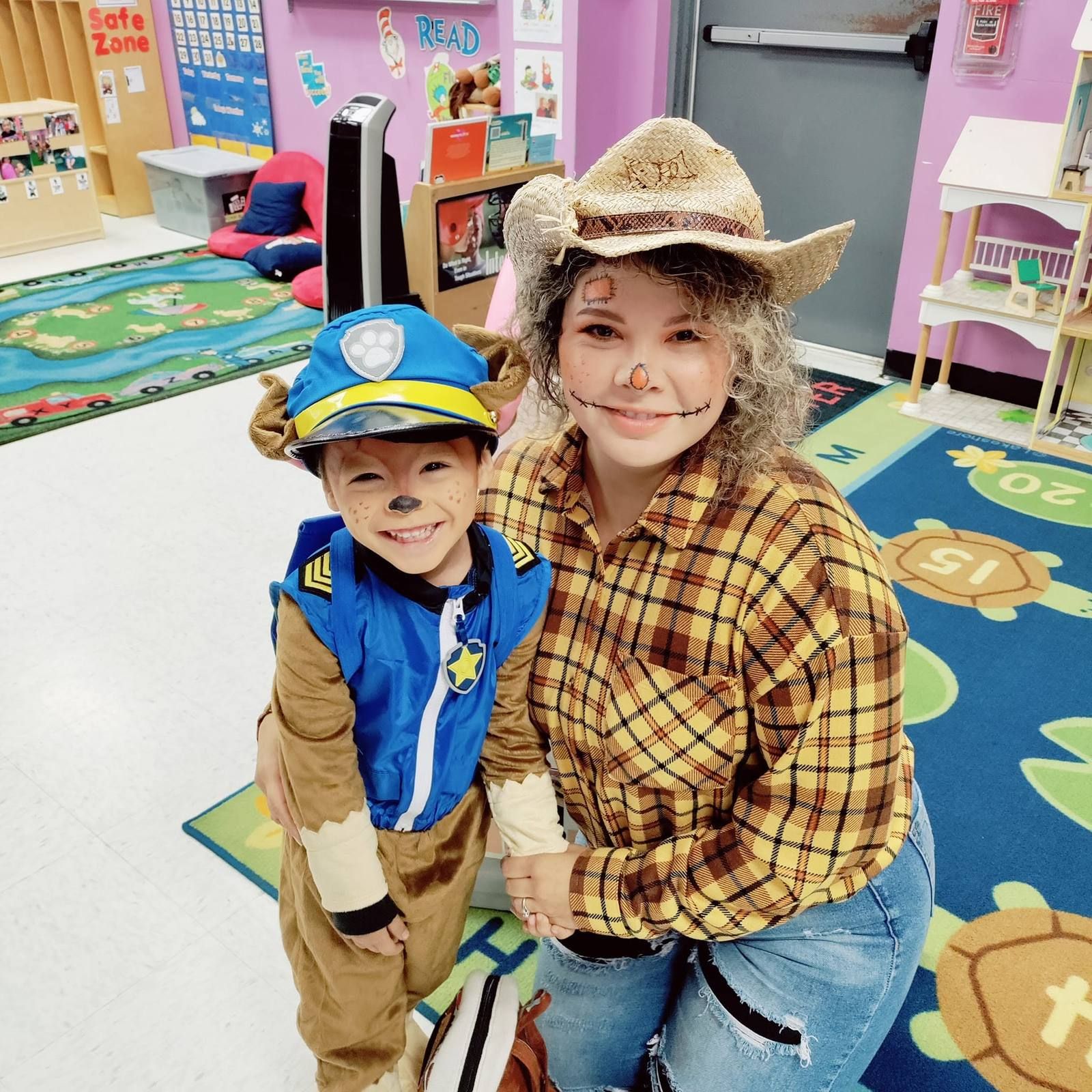 A teacher dressed as a scarecrow hugging a smiling toddler in a puppy costume