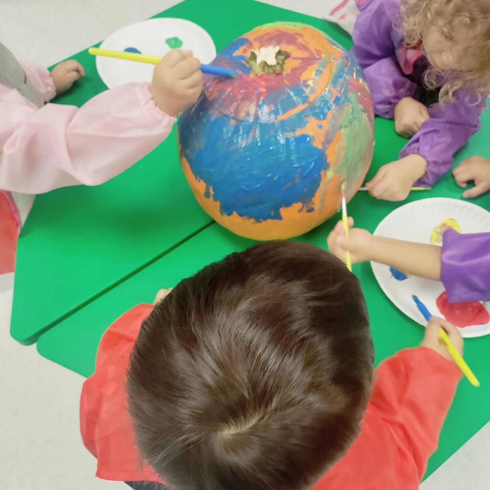 Children in paint smocks painting a big pumpkin together
