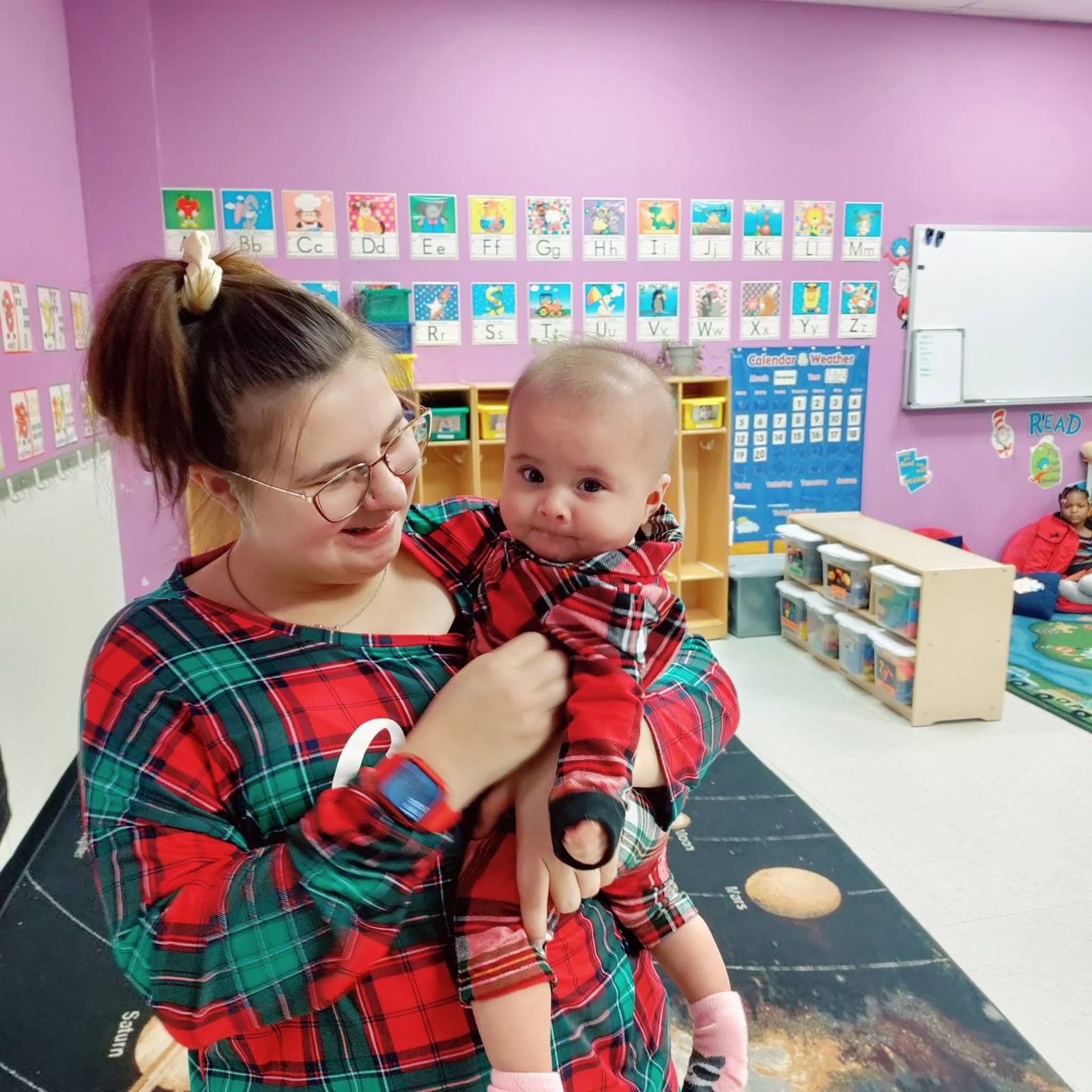 A teacher in holiday pajamas holding a baby in the classroom