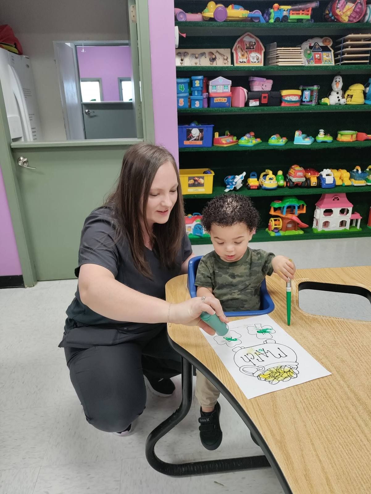 A teacher helping a toddler paint a shamrock picture