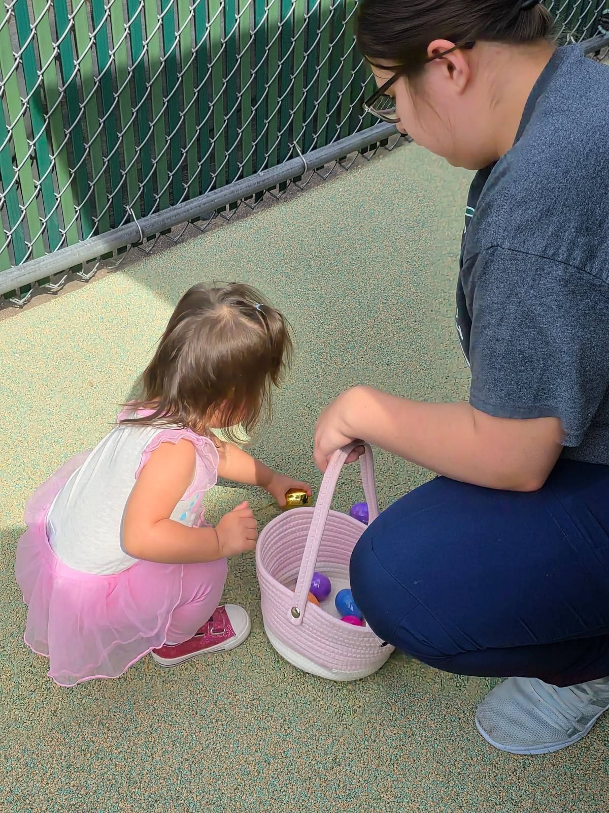 A teacher kneeling to help a toddler find eggs on the playground