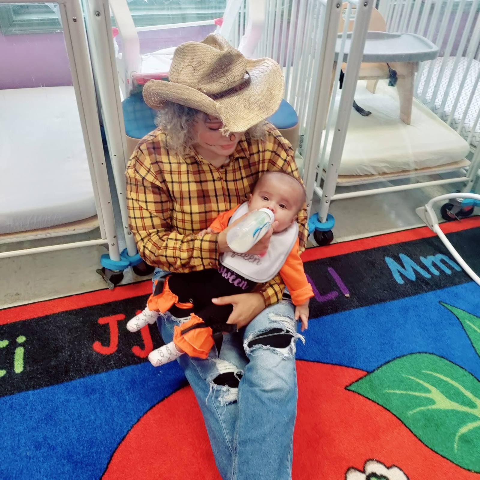 A teacher in a cowboy hat giving a baby a bottle on the rug