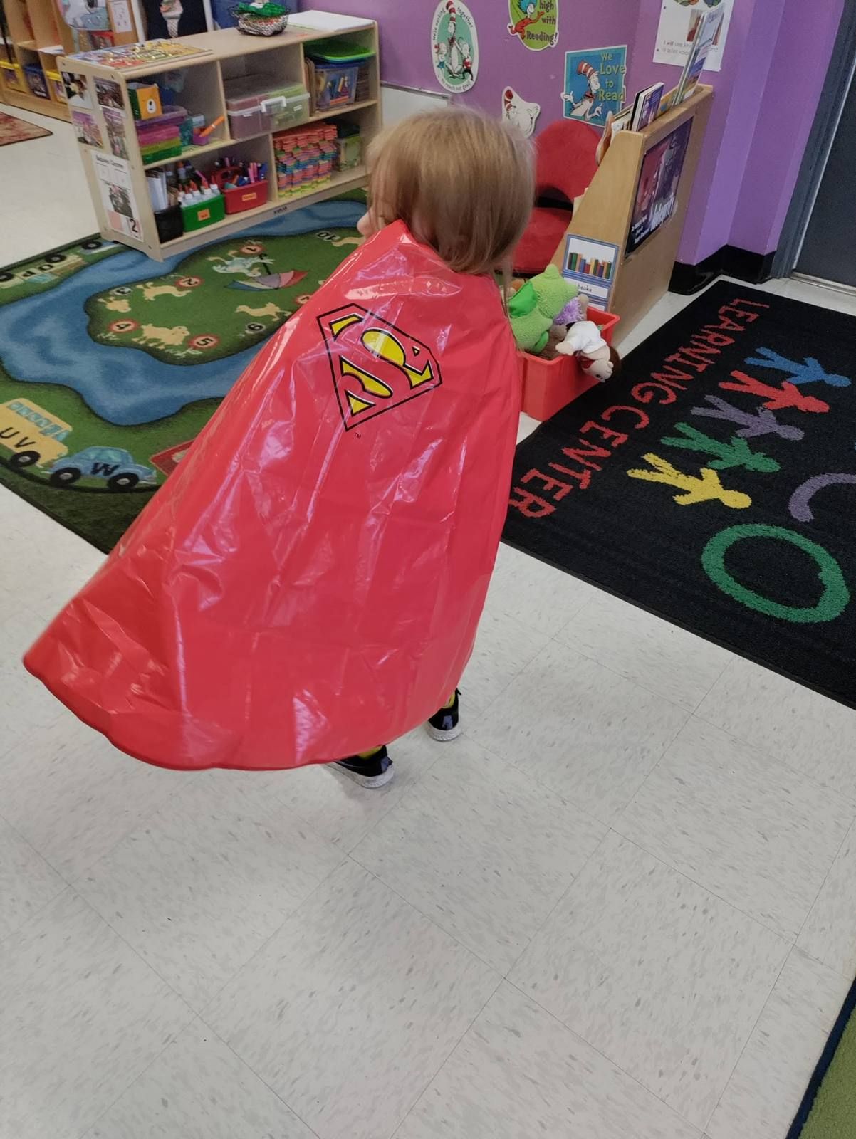 A toddler running in a red superhero cape